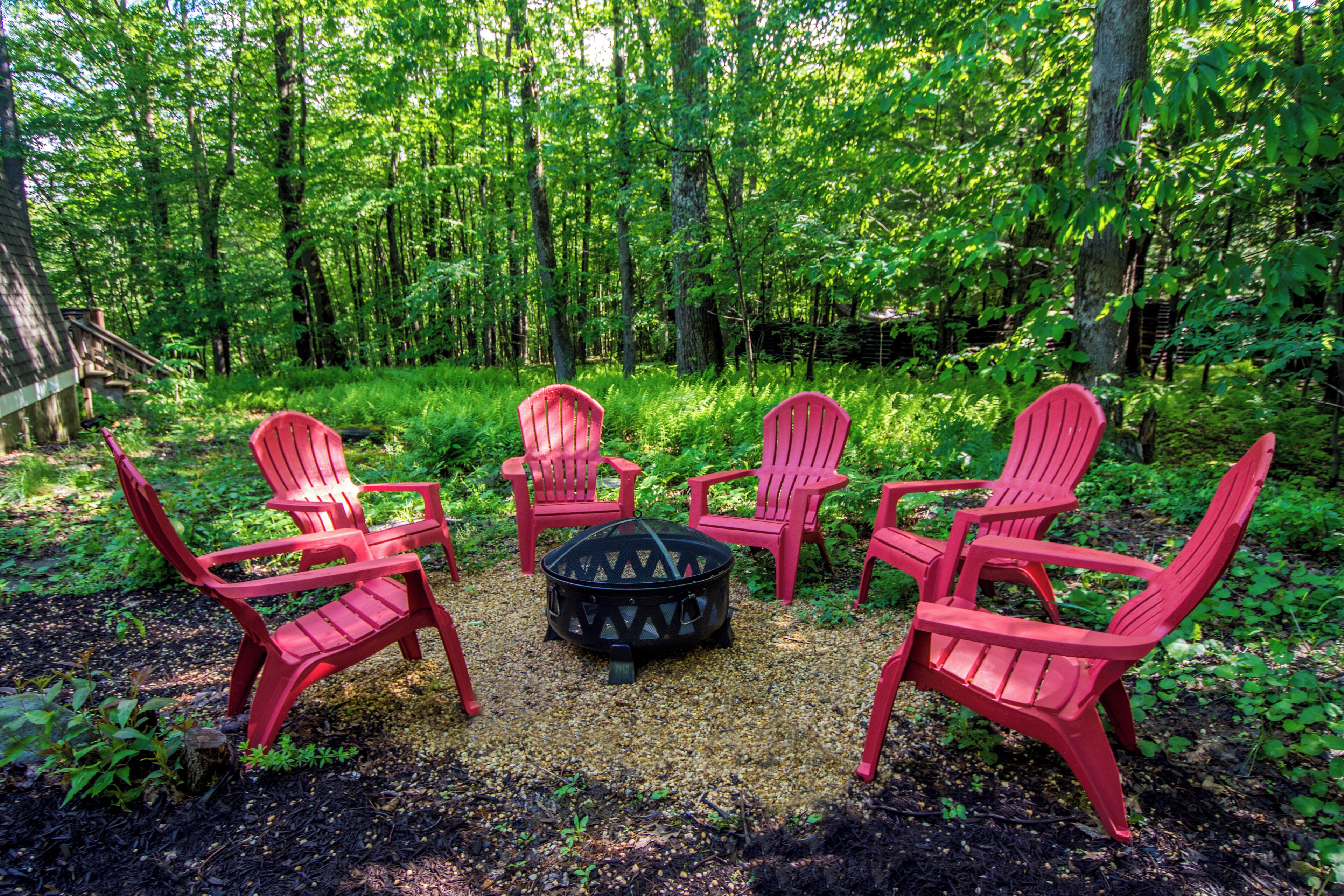 A circular fire pit surrounded by four red Adirondack chairs in a forested area.