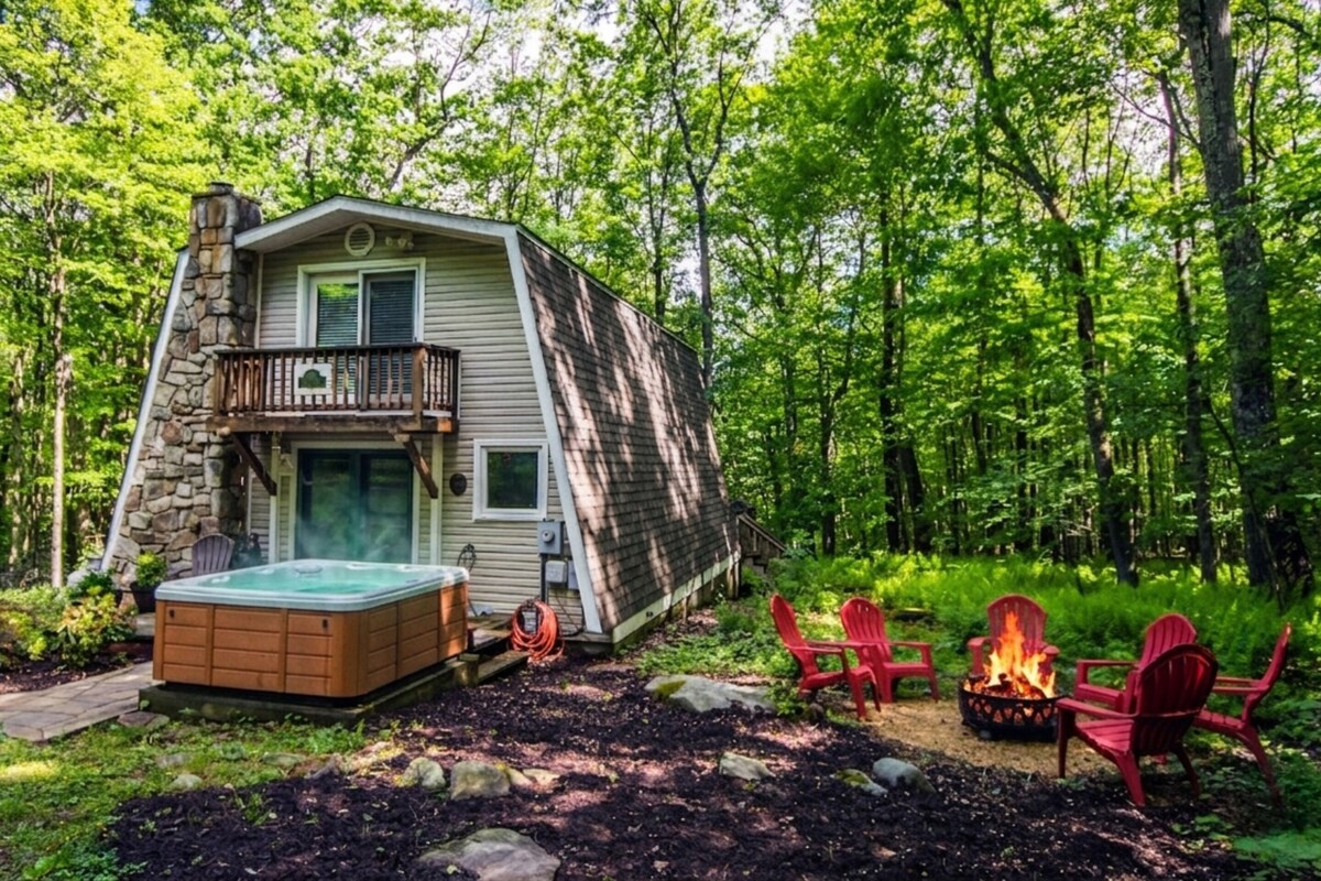 A cabin with a hot tub, outdoor seating, and a fire pit surrounded by trees.