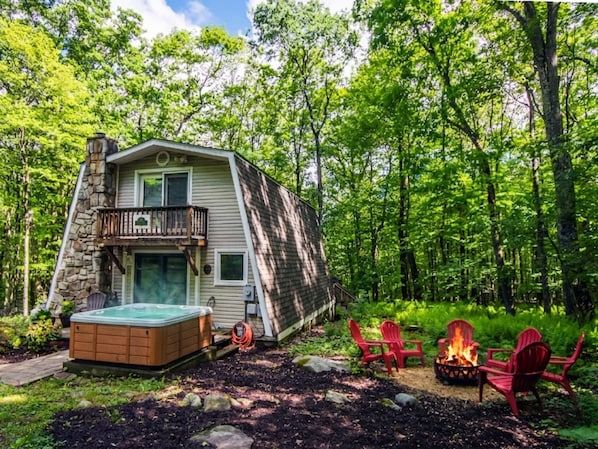 A cabin with a hot tub, outdoor seating, and a fire pit surrounded by trees.