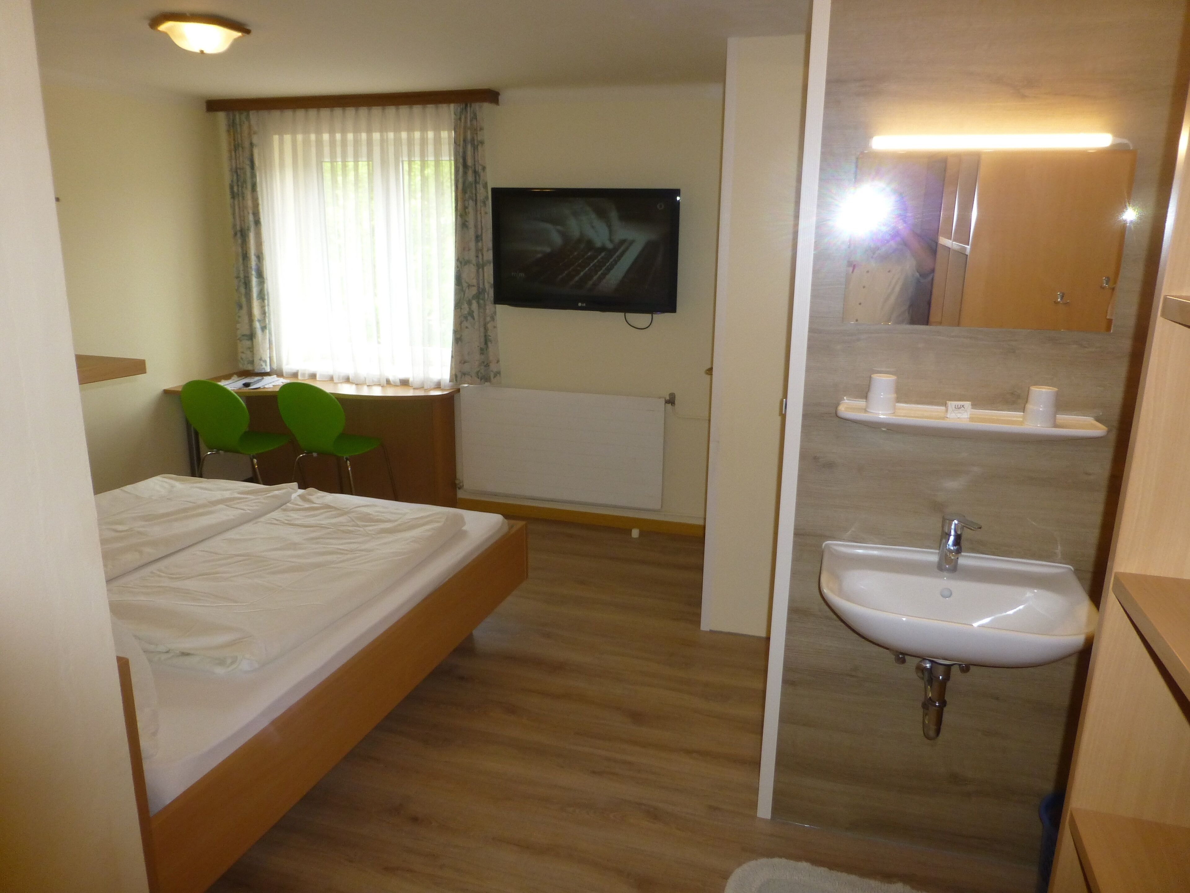 Habitación de hotel con cama, escritorio con silla, televisor, baño con lavabo y espejo.