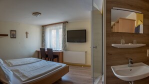 A hotel room with a bed, a desk with a chair, a television, a window with curtains, a radiator, a wooden cabinet, and a sink. - Smartmotel (Straß im Attergau)