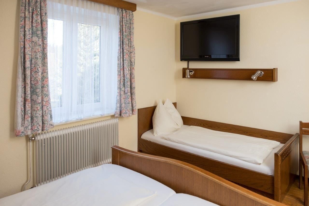 Habitación de hotel con una cama individual, una cama más pequeña, un televisor montado en la pared y una ventana con cortinas florales.