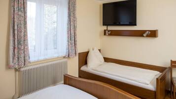 Habitación de hotel con una cama individual, una cama más pequeña, un televisor montado en la pared y una ventana con cortinas florales.