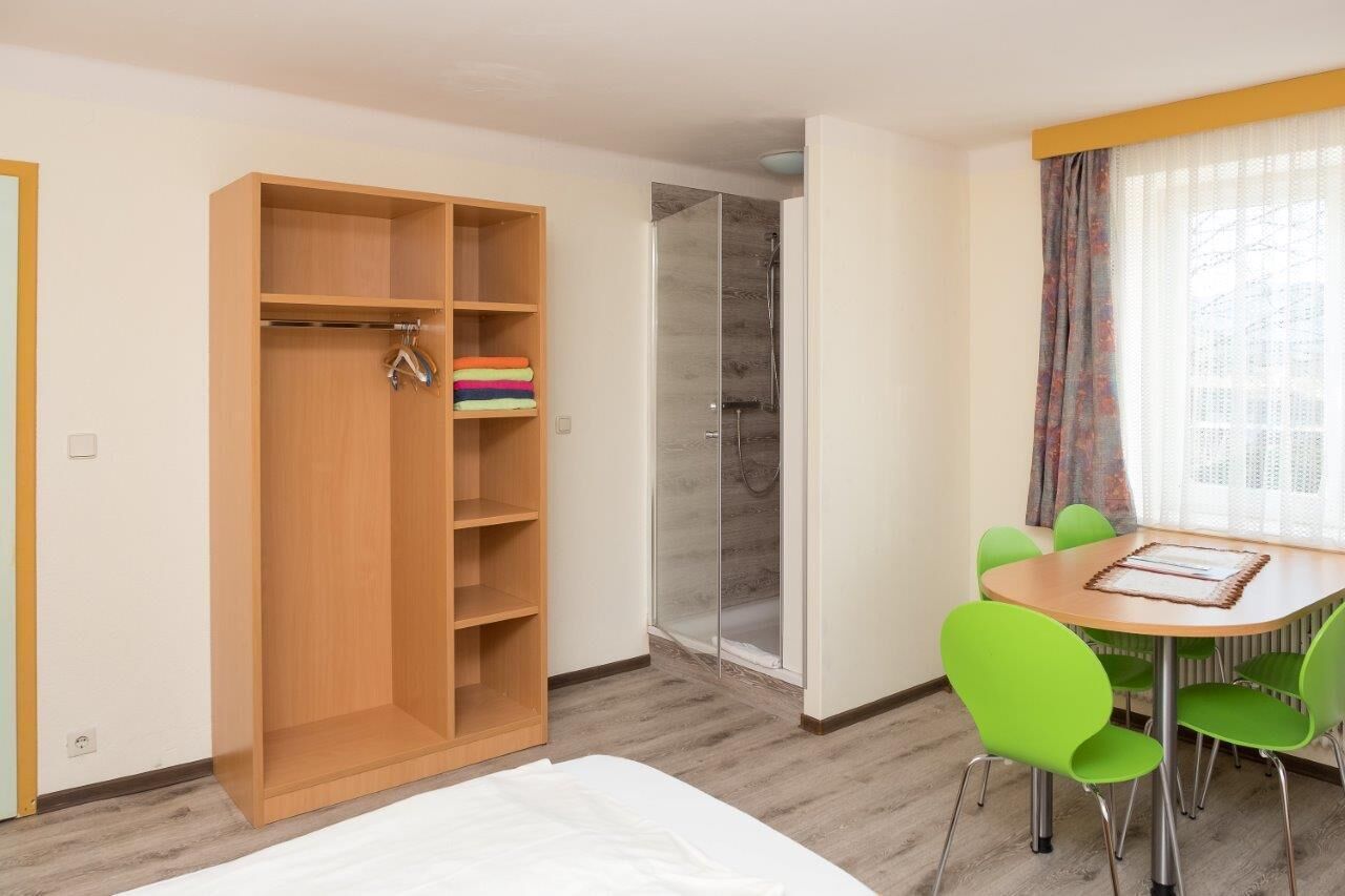 Ein Zimmer mit Holzschrank, Dusche, Esstisch mit grünen Stühlen und einem Vorhangfenster.