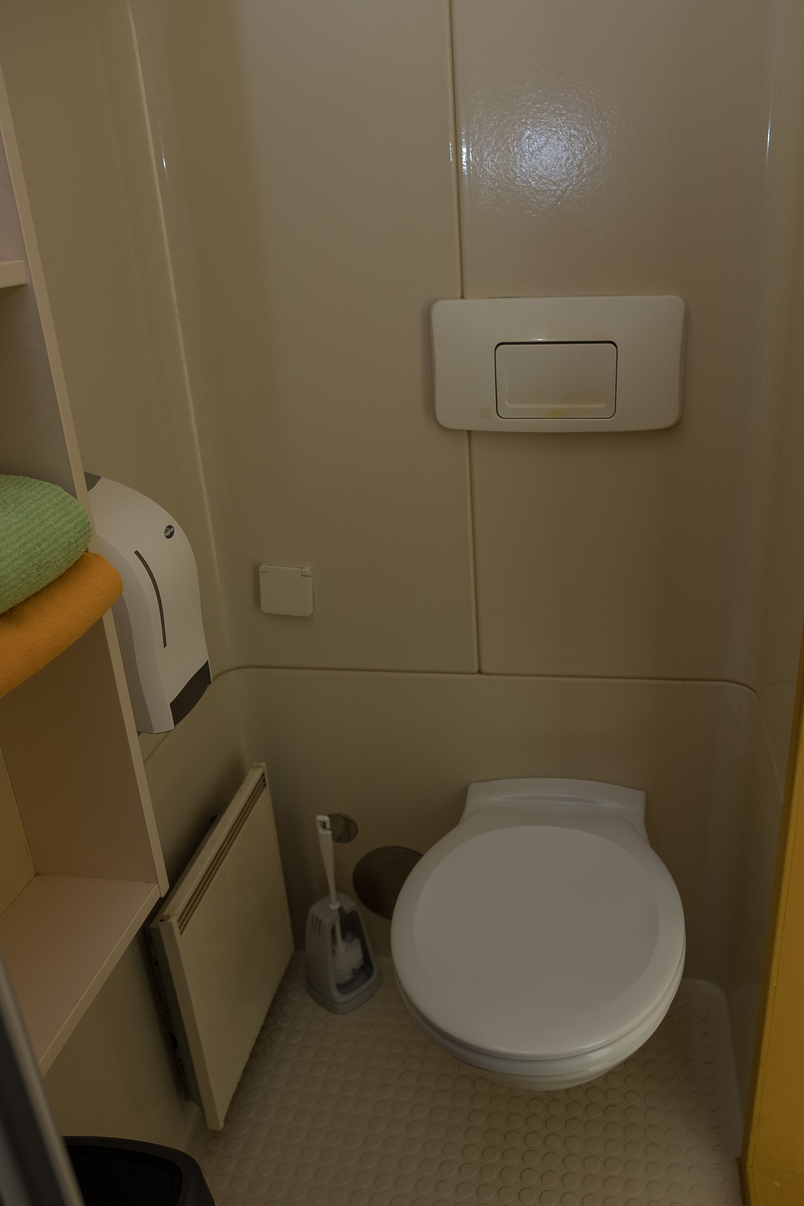 Une petite salle de bain compacte, équipée de toilettes, d’un sèche-mains, d’une poubelle et d’une étagère sur laquelle est posée une serviette verte.