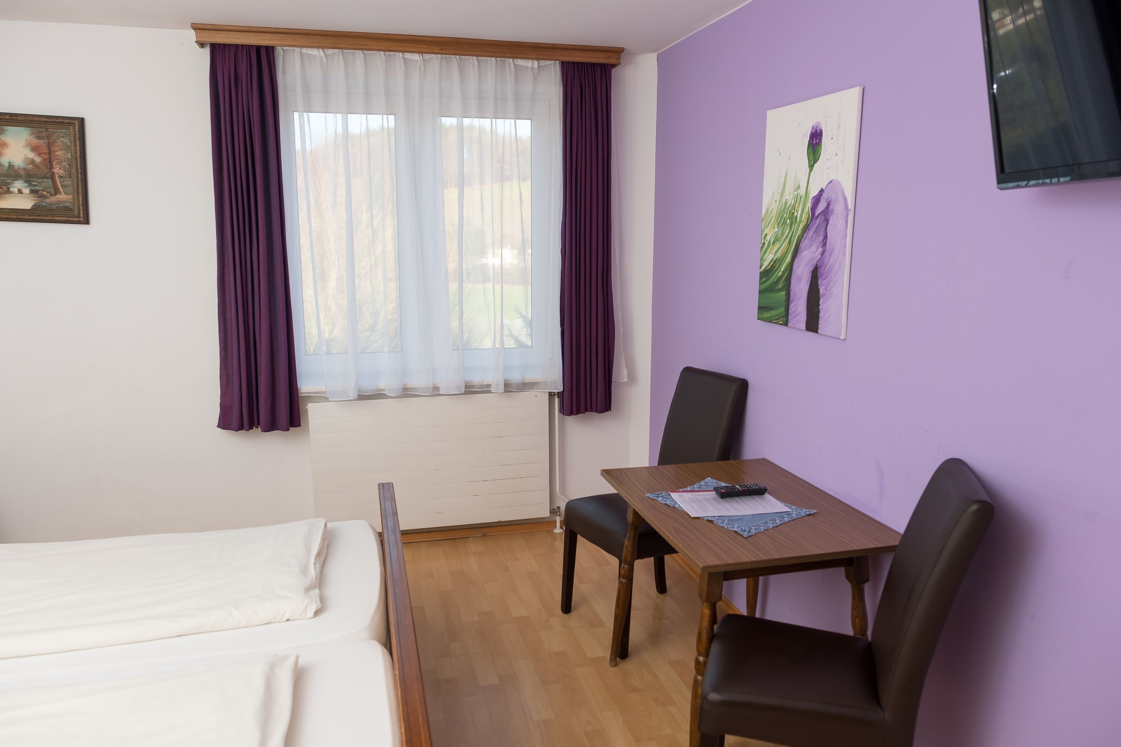 Een kamer met een bed, een tafel met stoel, een schilderij aan de muur en een raam met gordijnen.