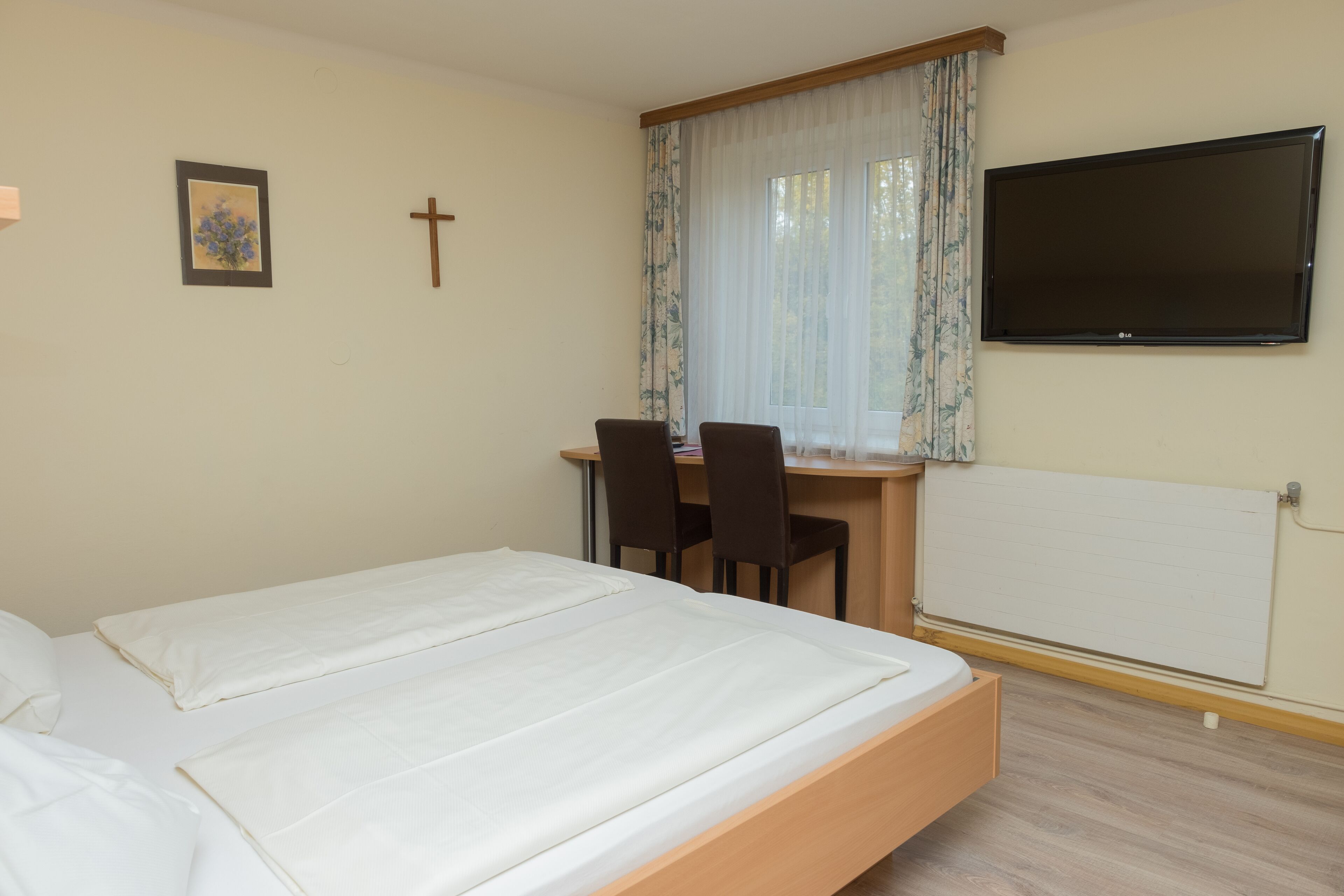 A bedroom with a bed, a desk with two chairs, a TV, and a window with curtains.