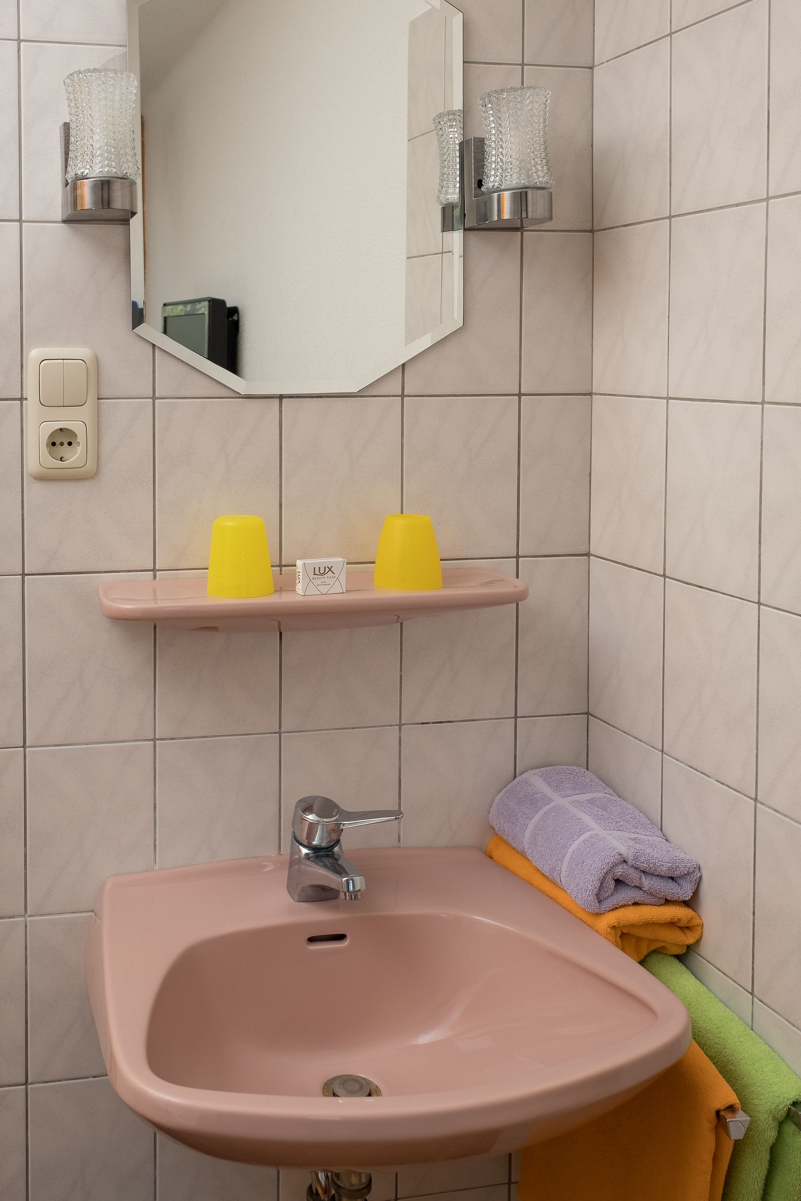 Ein Badezimmer mit einem rosafarbenen Waschbecken, einem Spiegel und einem Regal mit gelben Tassen sowie einer Lux-Seifenspender.