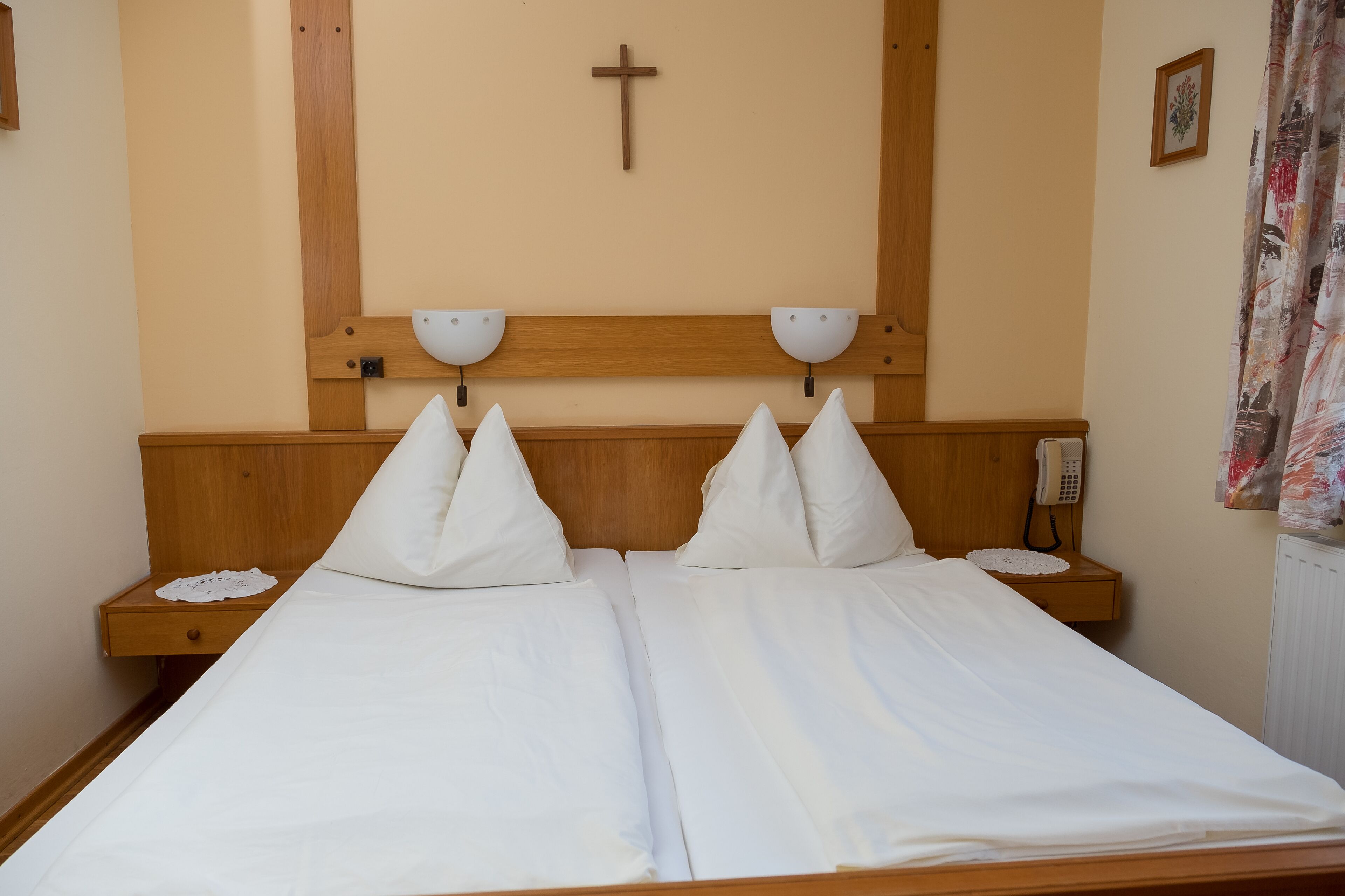 A double bed with white linens, two bedside tables with lamps, a wooden headboard, a cross on the wall, and a telephone on the nightstand.