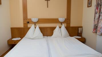 A double bed with white linens, two bedside tables with lamps, a wooden headboard, a cross on the wall, and a telephone on the nightstand.