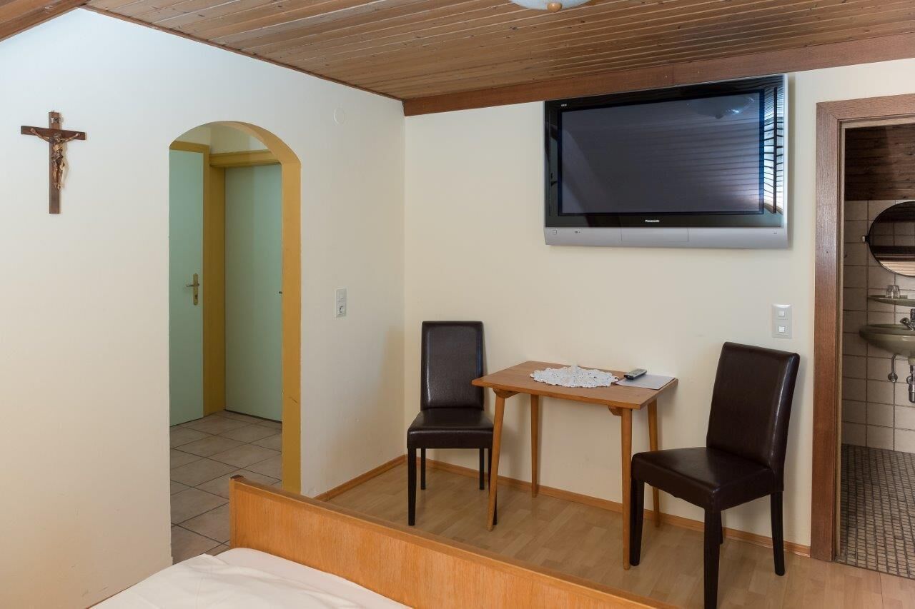 Una habitación con cama, televisor montado en la pared, dos sillones, una mesita y una cruz en la pared.