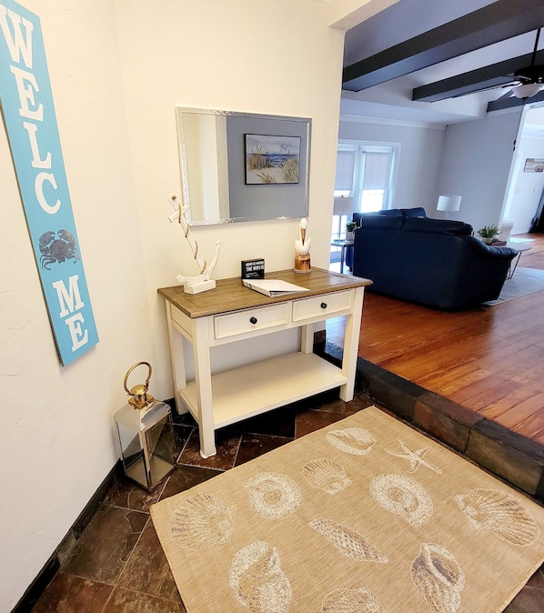 Ein Wohnzimmer mit einem weißen Konsolentisch, einem gerahmten Bild darüber, einem blauen 'WELCOME'-Schild, einem dunklen Sofa und einem gemusterten Teppich.