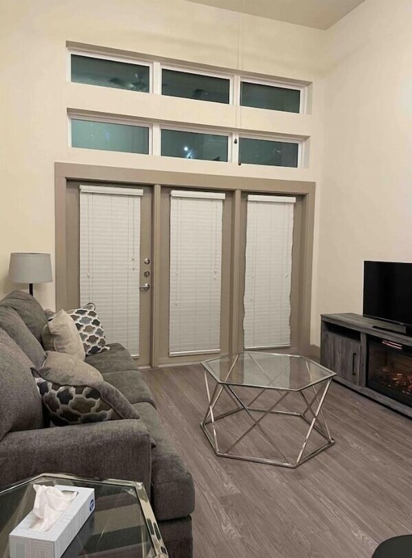 Una sala de estar con un sofá, una mesa de centro de vidrio y un televisor. Hay dos puertas correderas con persianas.