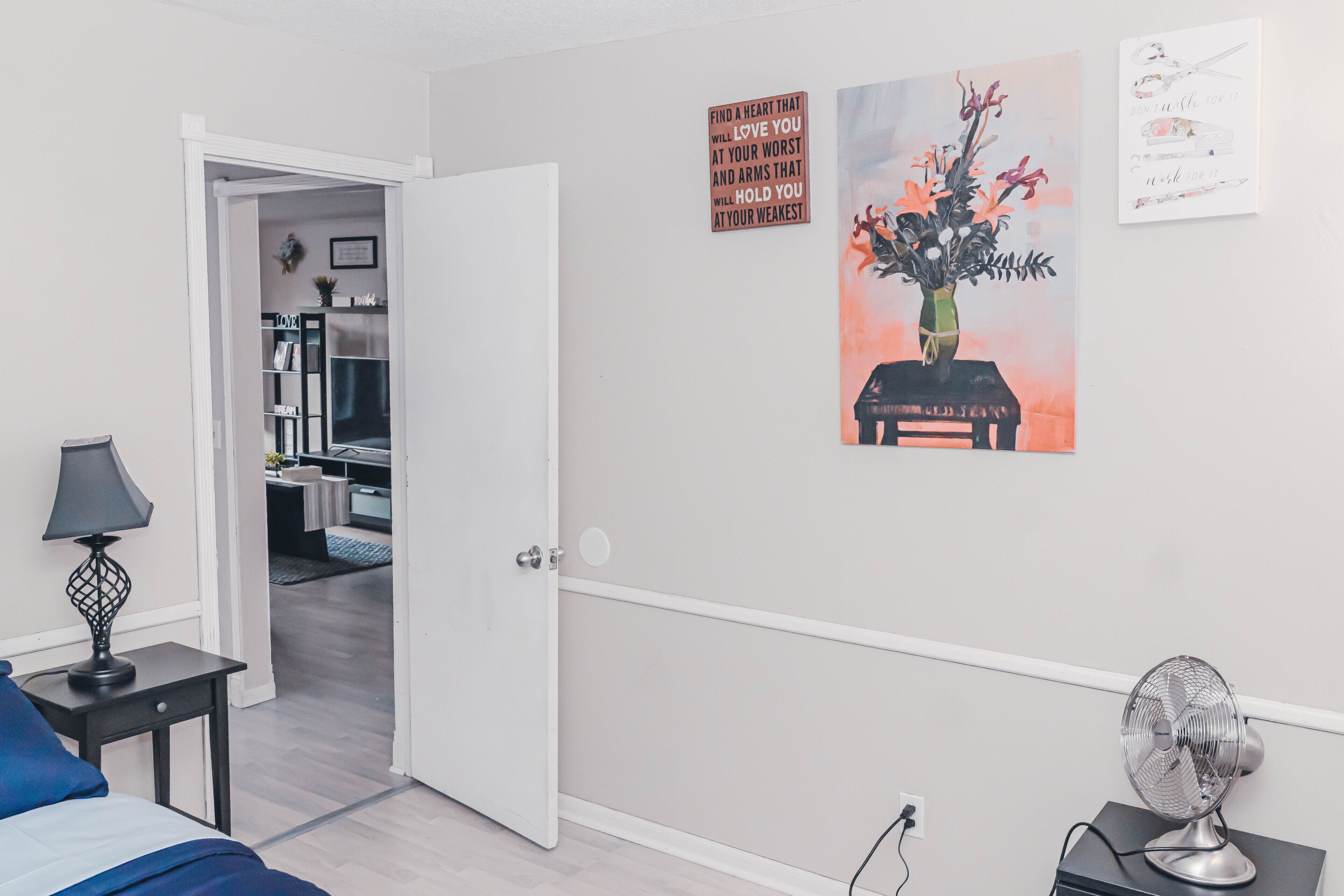 A bedroom with a bed, a nightstand, a lamp, a wall-mounted fan, and a door leading to another room.