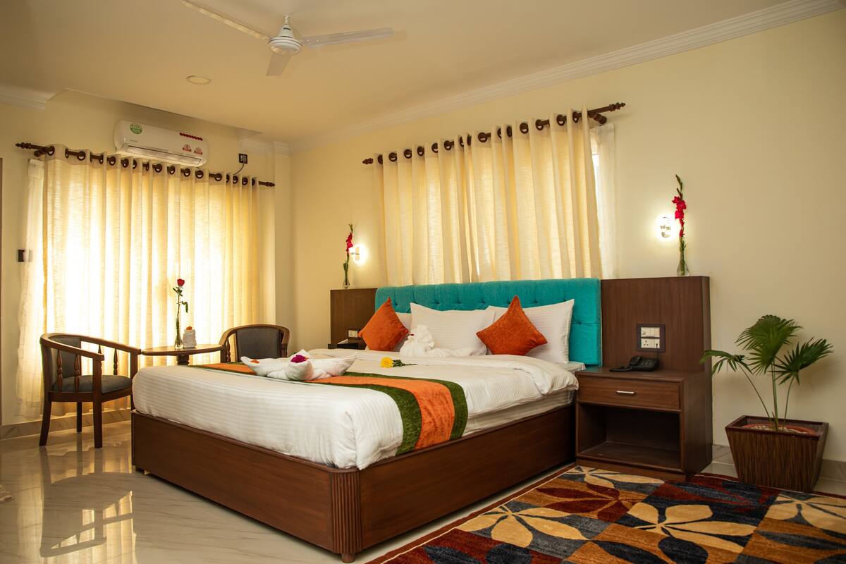A hotel room with a bed, a desk, a chair, a nightstand, a plant, and an air conditioning unit.