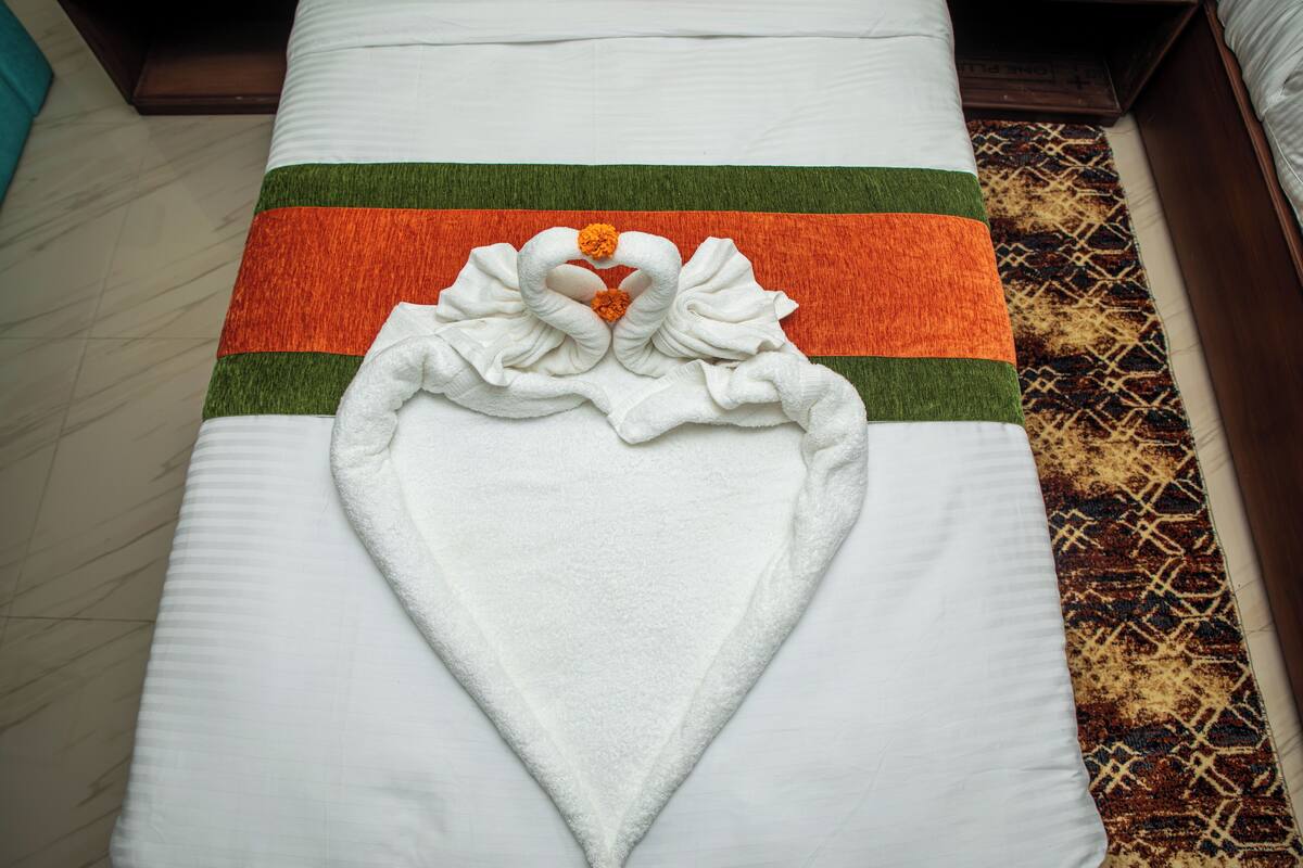 A neatly made bed with a towel arranged in the shape of a heart and two swans, a green and orange striped pillow, and a patterned rug.
