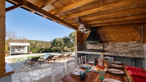 An outdoor dining area with a wooden table set for a meal, surrounded by chairs, and a view of a swimming pool and greenery in the background. - Villa Serenity (Corfu)