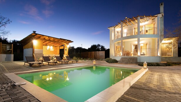 A swimming pool with lounge chairs and a covered seating area, surrounded by a brick patio and a two-story building with balconies. - Villa Serenity (Corfu)