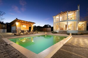 A swimming pool with lounge chairs and a covered seating area, surrounded by a brick patio and a two-story building with balconies. - Villa Serenity (Corfu)