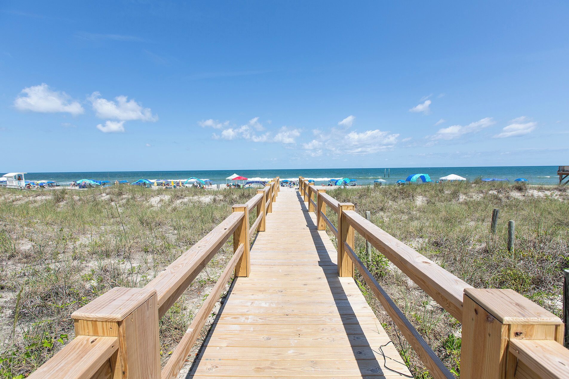 A wooden boardwalk leading to a beach with umbrellas and beachgoers.