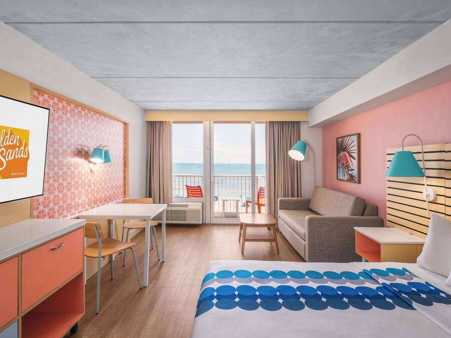 Una camera d'albergo con un letto, una scrivania, una sedia, un divano, una TV e un balcone vista mare.