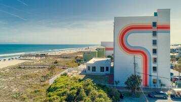 A coastal building with a distinctive red and beige curved design on its facade, situated near a beach with a wooden pier and beachgoers.