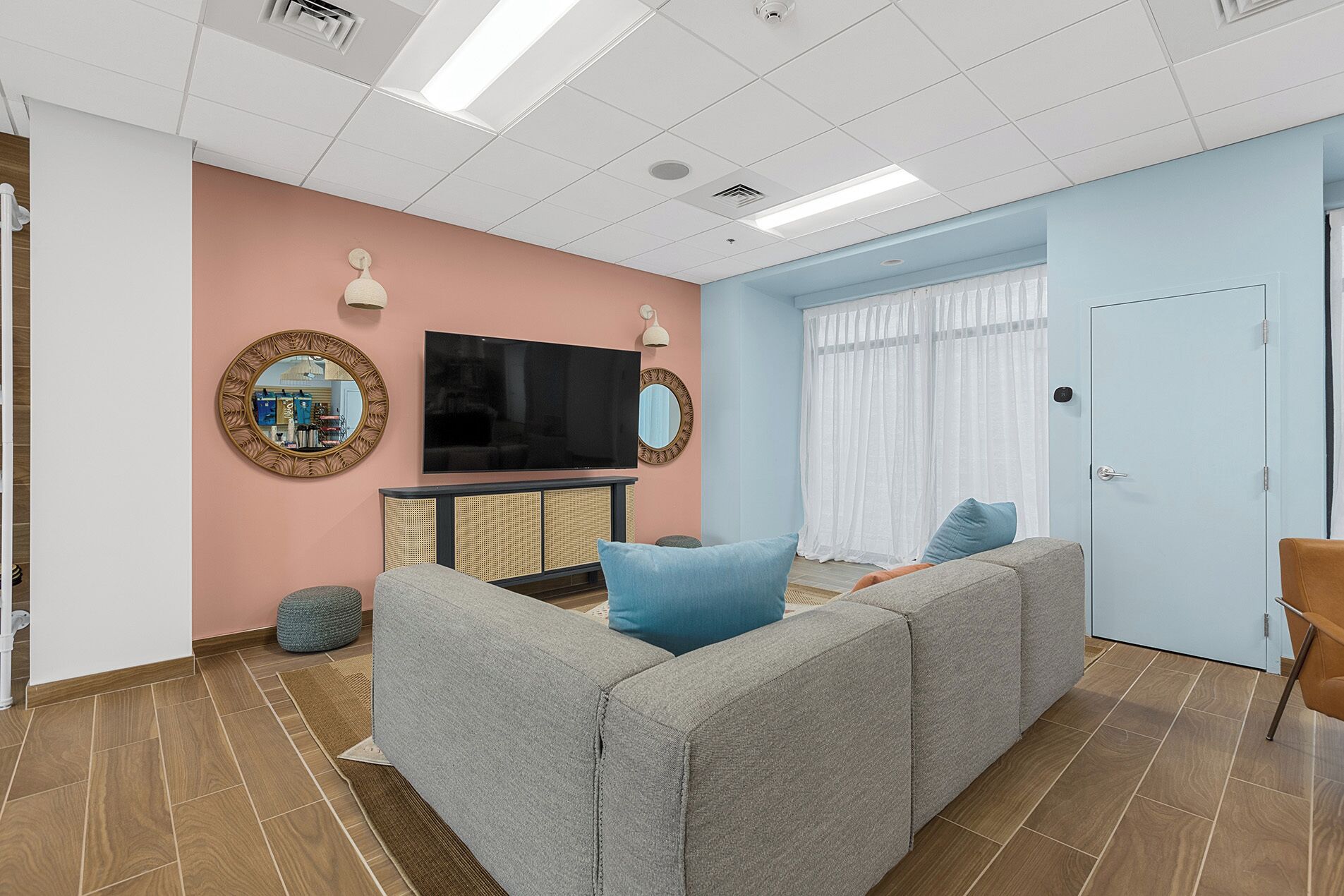 Ein Wohnzimmer mit einem großen Flachbildfernseher, zwei Wandspiegeln, einem grauen Ecksofa und einer hellblauen Tür.