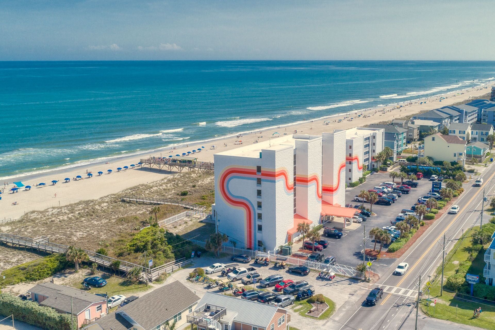 A coastal hotel with a distinctive red and white facade, a parking lot, and a beach view.