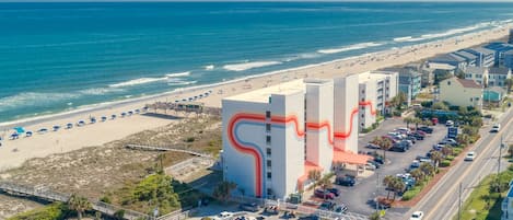 A coastal hotel with a distinctive red and white facade, a parking lot, and a beach view.