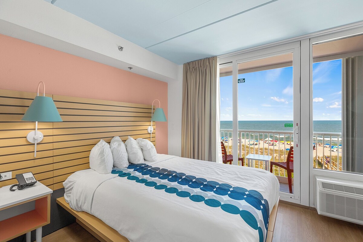 A hotel room with a bed, a desk with a phone, a window with a view of the beach, and a balcony with a table and chairs.