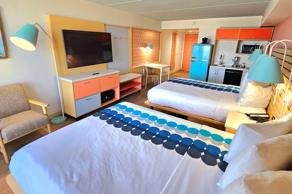 A hotel room with a large bed, a desk, a television, a kitchenette, and a chair.