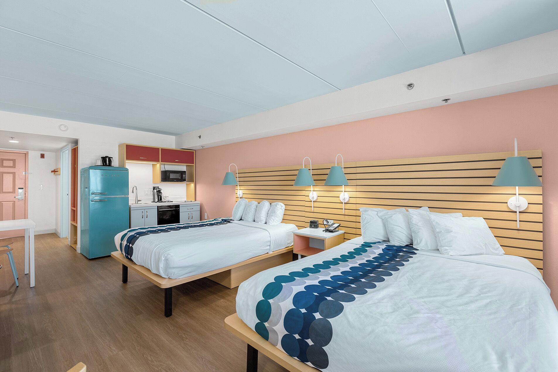 Une chambre d’hôtel avec deux lits, une kitchenette et une tête de lit en bois agrémentée de lampes fixées au mur.