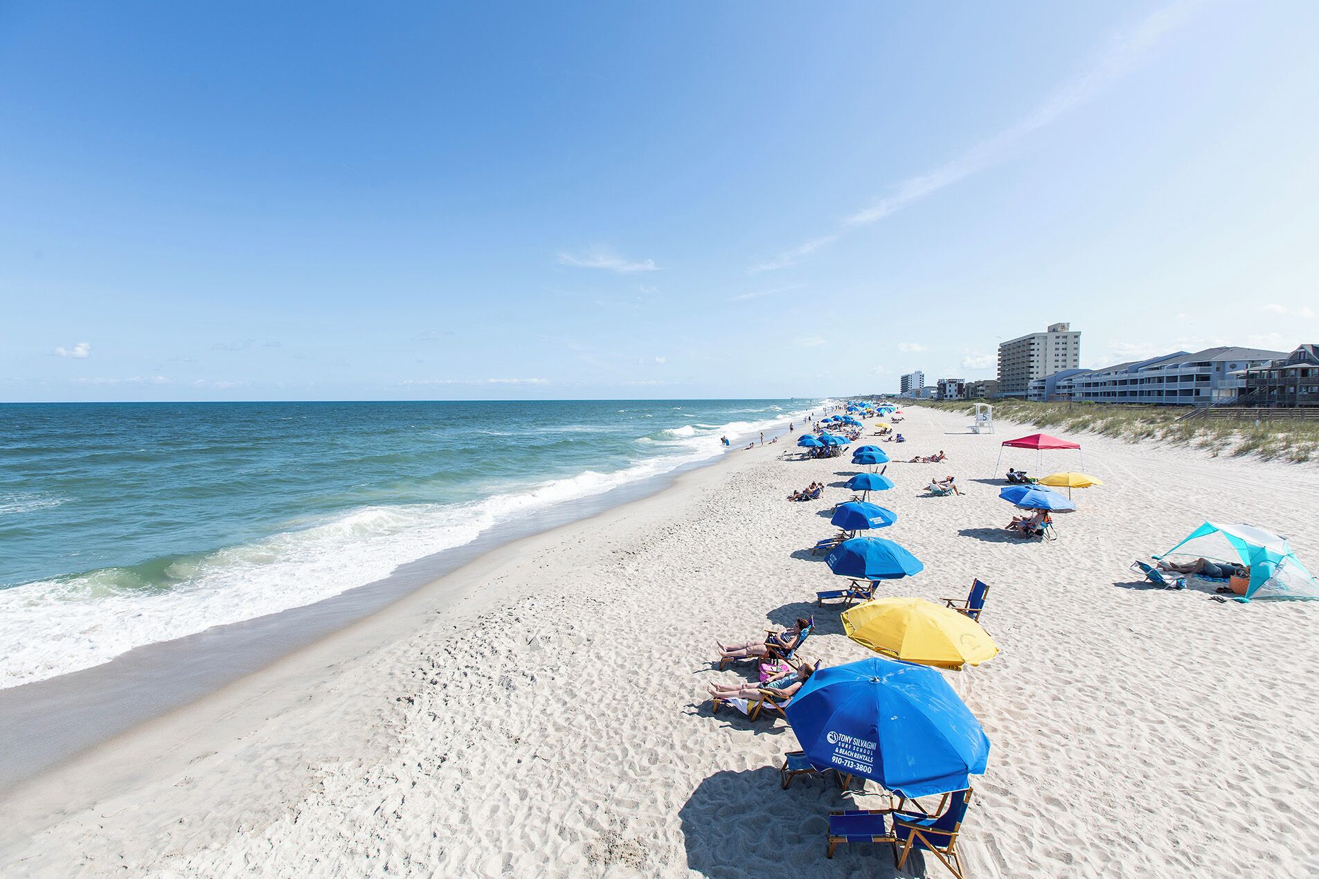 A beach with rows of beach chairs and umbrellas, a clear sky, and buildings in the background.