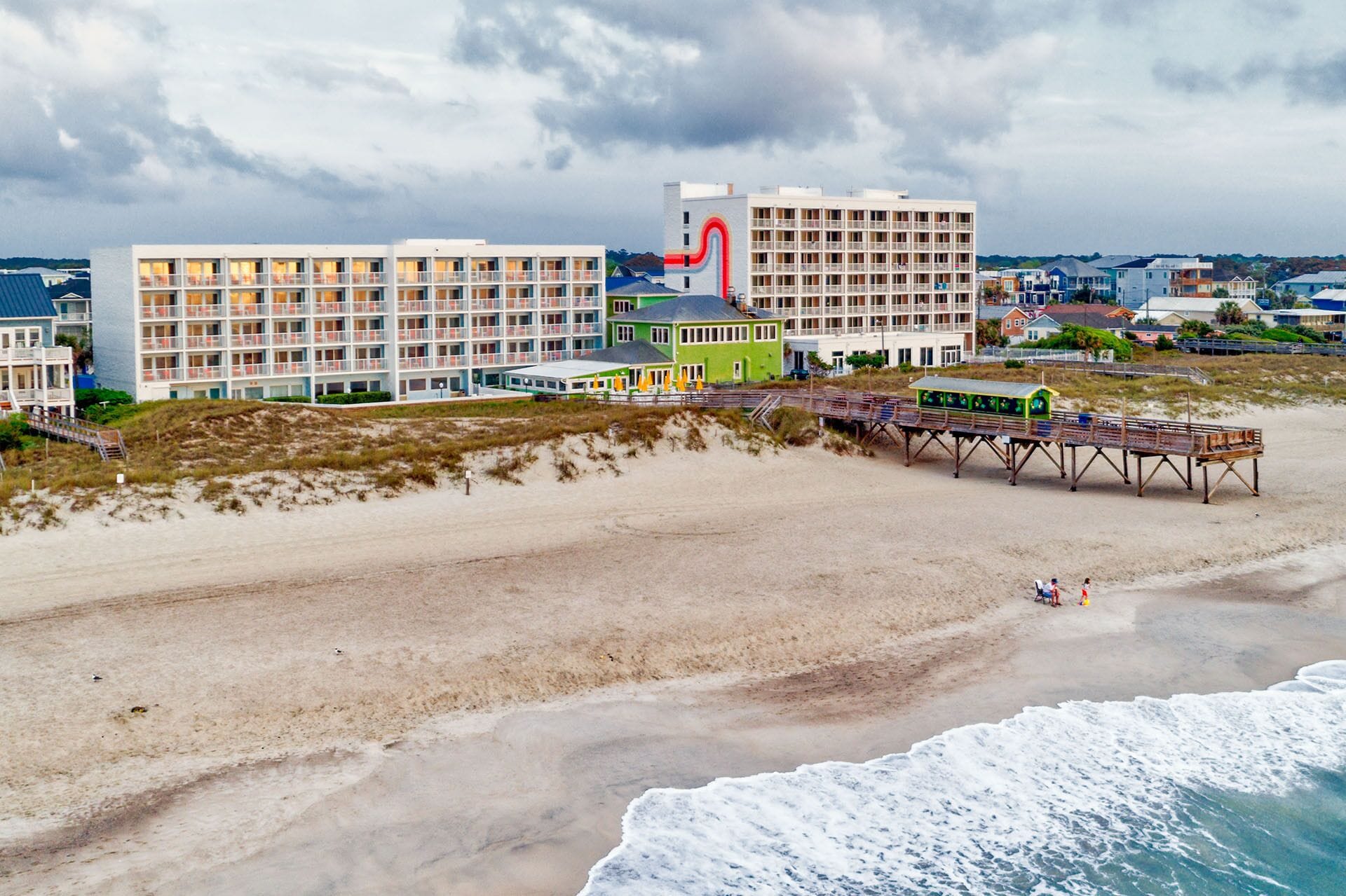 A coastal hotel with multiple floors, a beach view, and a pier with a green building.