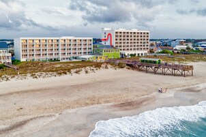 A coastal hotel with multiple floors, a beach view, and a pier with a green building.