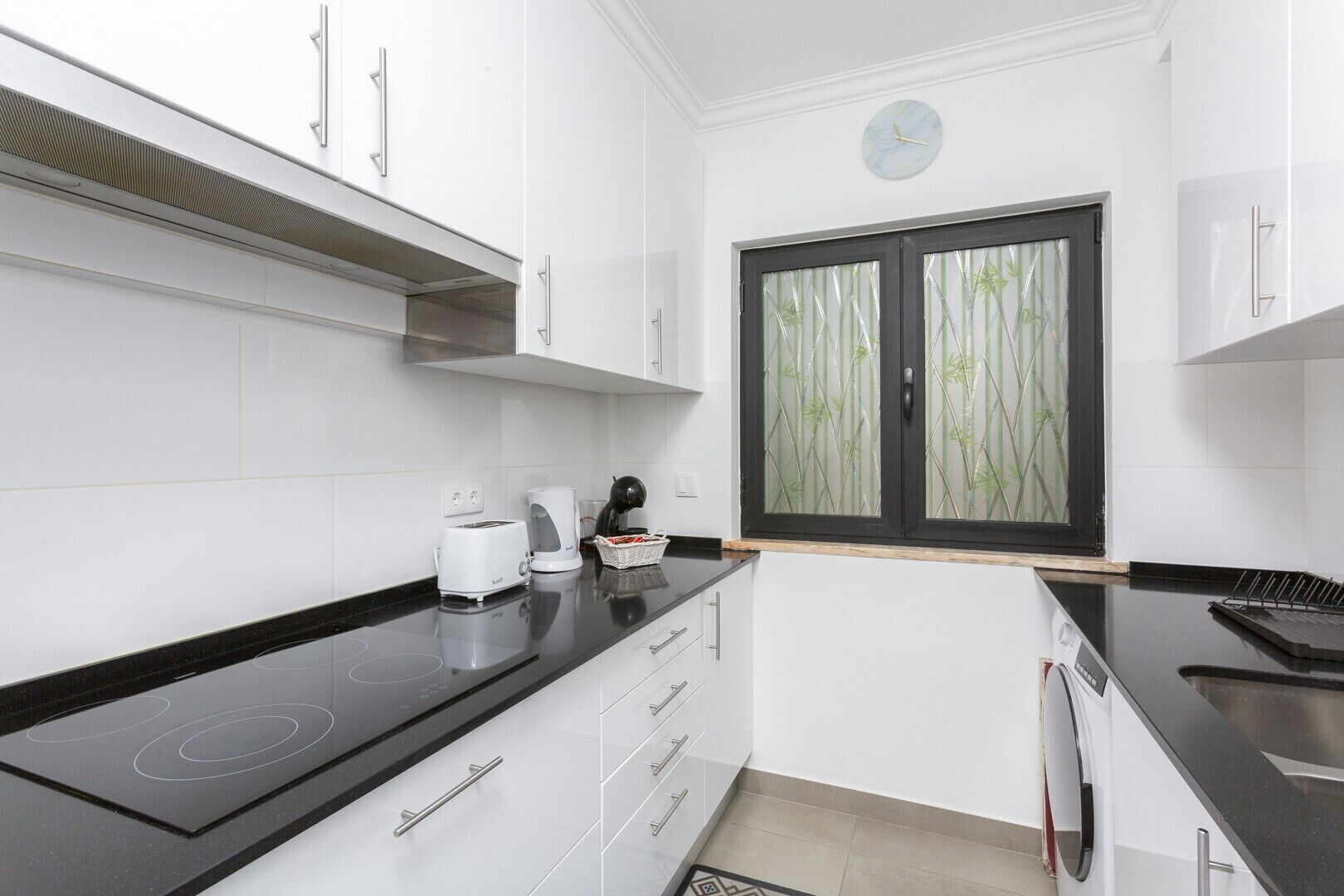 A modern kitchen with white cabinets, a black countertop, a toaster, a kettle, and a window with bamboo-patterned blinds.