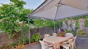 Een terras buiten met een tafel en stoelen onder een luifel, omringd door groen en een stenen muur.