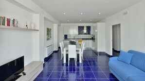 Een moderne woonkamer met een blauw betegelde vloer, een witte eettafel met stoelen, een blauwe bank en een flatscreen televisie.