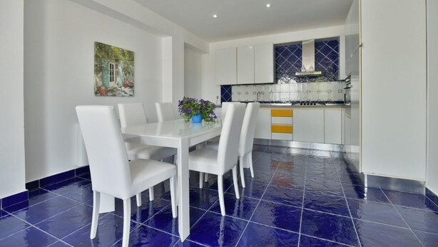 Een eethoek met een witte tafel en stoelen, een vloer van blauwe tegels, en een keuken achtergrond met witte kasten en een blauwe achterwand.