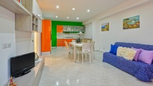 Uma sala de estar moderna com televisão de tela plana, uma área de jantar com mesa e cadeiras, uma cozinha com armários laranjas e um sofá azul com almofadas decorativas.