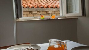 A table with a bowl of croissants and a pitcher of orange juice, a window with a view of a stone wall and a tiled roof, and a partially open window blind. - PK Apartments - Dubrovnik (Dubrovnik)