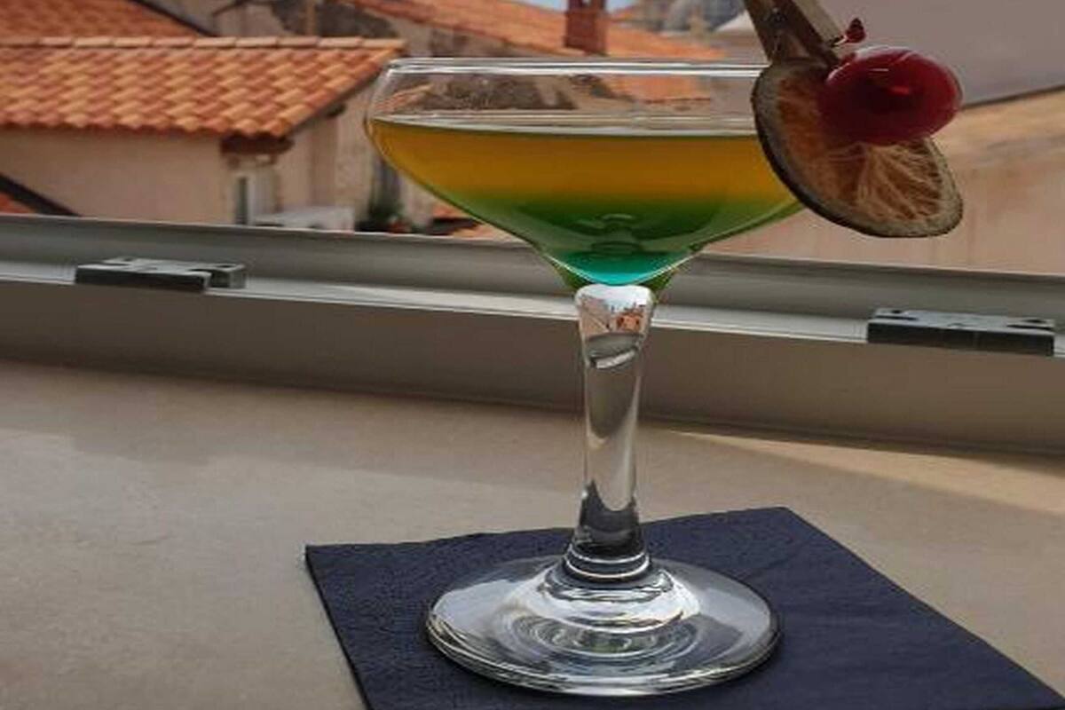 Un verre à cocktail contenant un liquide vert et orné d’une cerise sur le bord, posé sur une serviette sombre près d’une fenêtre donnant sur des bâtiments aux toits recouverts de tuiles rouges.
