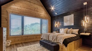 Een slaapkamer met een schuine kap, houten wanden, een groot bed met een zwart hoofdeinde, een bankje, een zacht, wit vloerkleed en een nachtkastje met een plant.