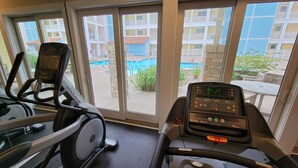 Una cinta de correr y una máquina elíptica en una habitación con ventanas amplias que dan a una piscina y un edificio con balcones.