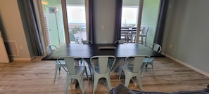 A dining area with a large table and six chairs, a view of the beach through a glass door, and a wooden floor. - Sandbridge 3bd with stunning Bay views!  Beach front complex! Newly remodeled! (Virginia Beach)