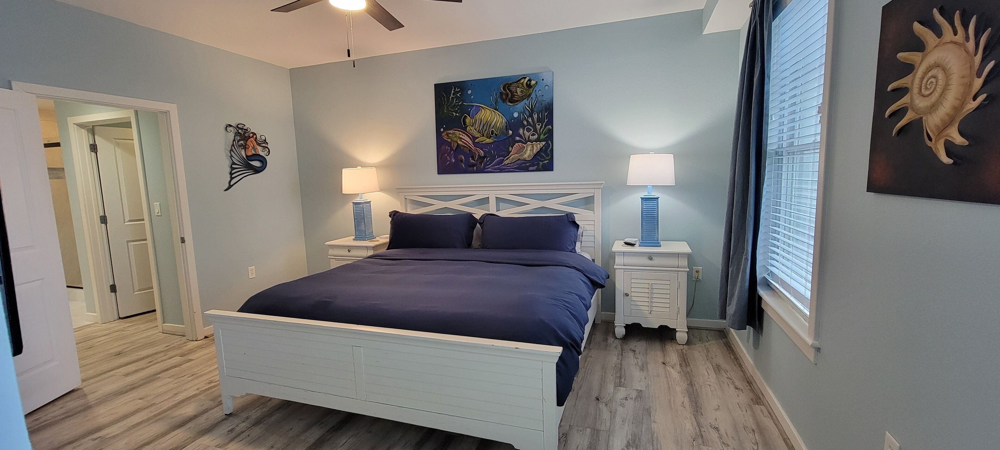 Un dormitorio con cama, dos mesitas de noche, ventilador de techo y obras de arte en las paredes.