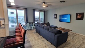 Una sala moderna con un sofá gris, una mesa de centro y un televisor de pantalla plana montado en la pared. Se ve un balcón con comedor a través de puertas de vidrio.