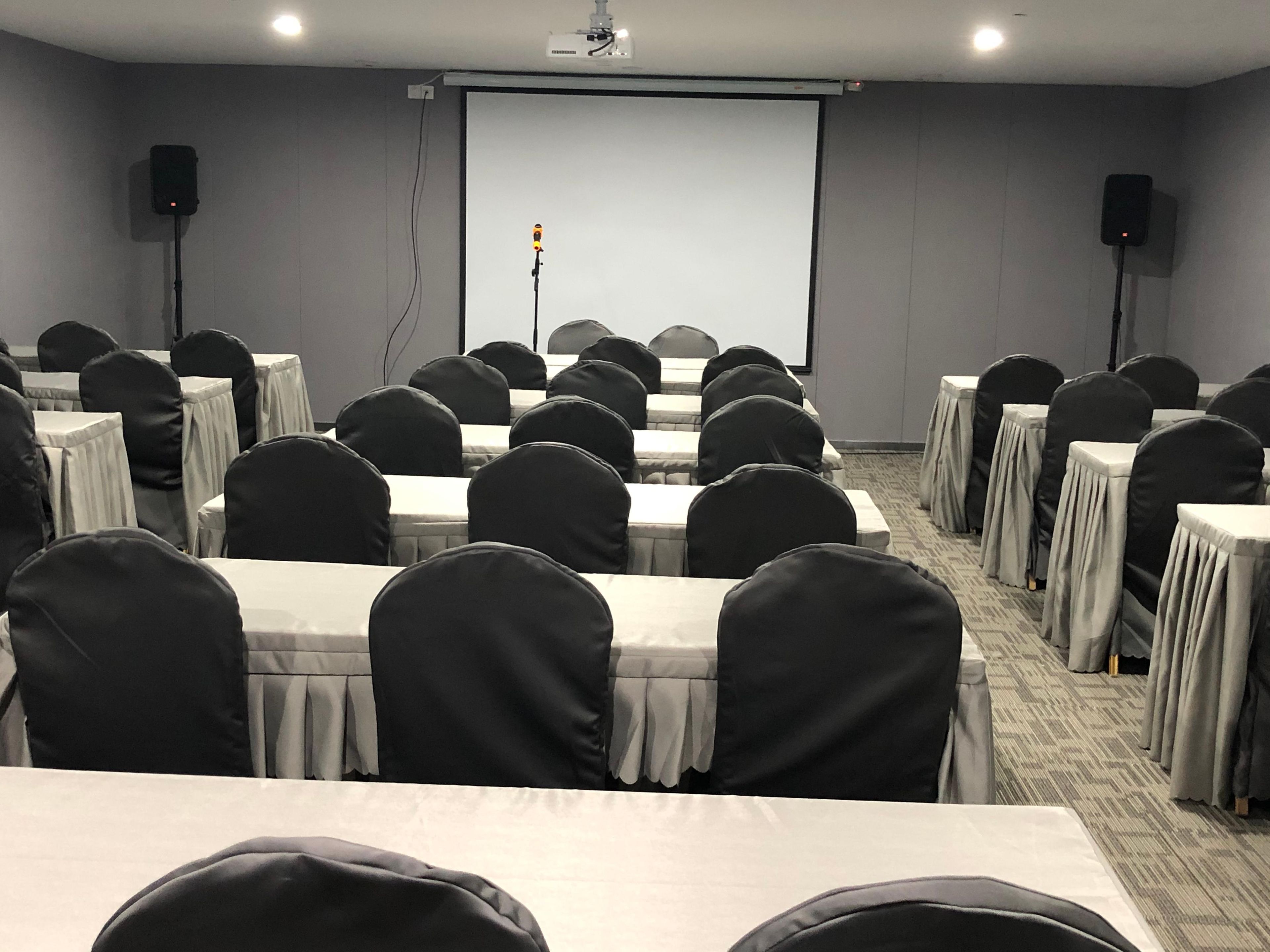 A conference room with rows of tables and chairs, a projector screen, and speakers.