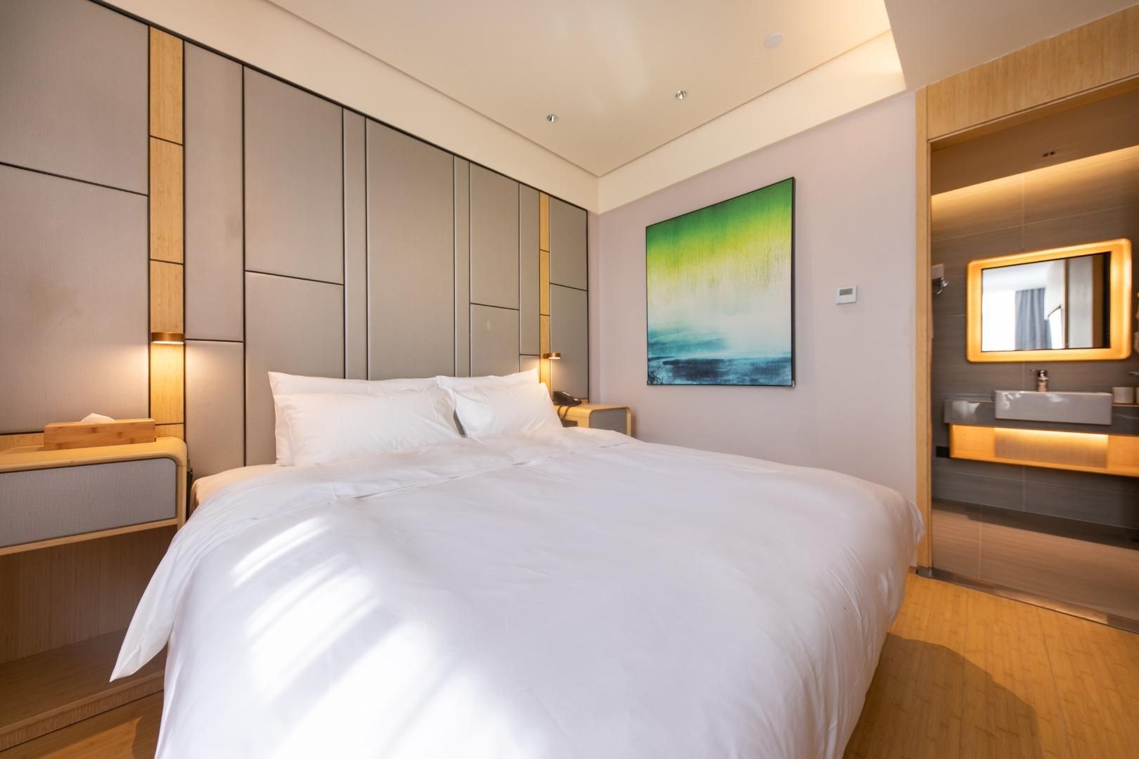 Una habitación de hotel moderna con una cama grande, una mesita de noche, un tocador, un cuadro en la pared y un baño con espejo y lavamanos.