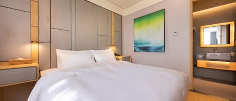 A modern hotel room with a large bed, a bedside table, a nightstand, a wall-mounted painting, and a bathroom with a mirror and sink.